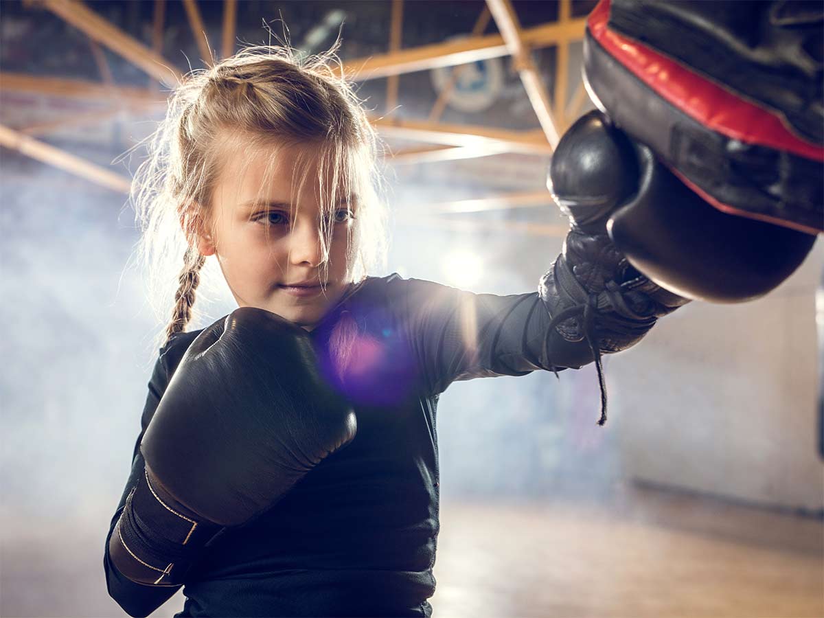 A young girl wearing boxing gloves punches an instructor's glove while training kickboxing.
