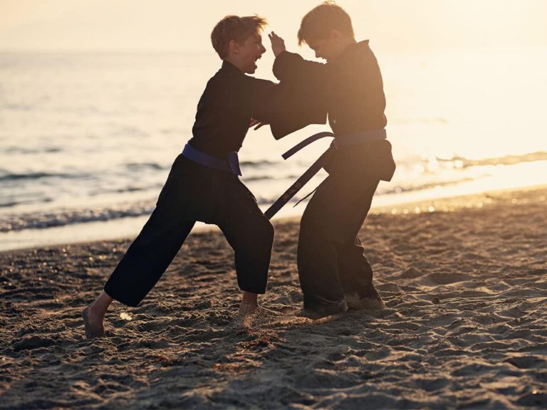 Two kids in black robes engage in martial arts together on the shore of a beach.