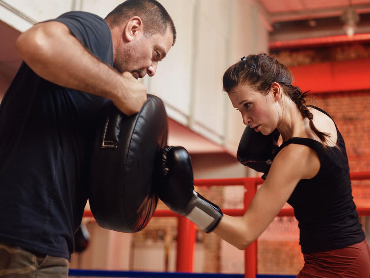A woman trains for kickboxing by uppercutting a safety cushion held by a male trainer.